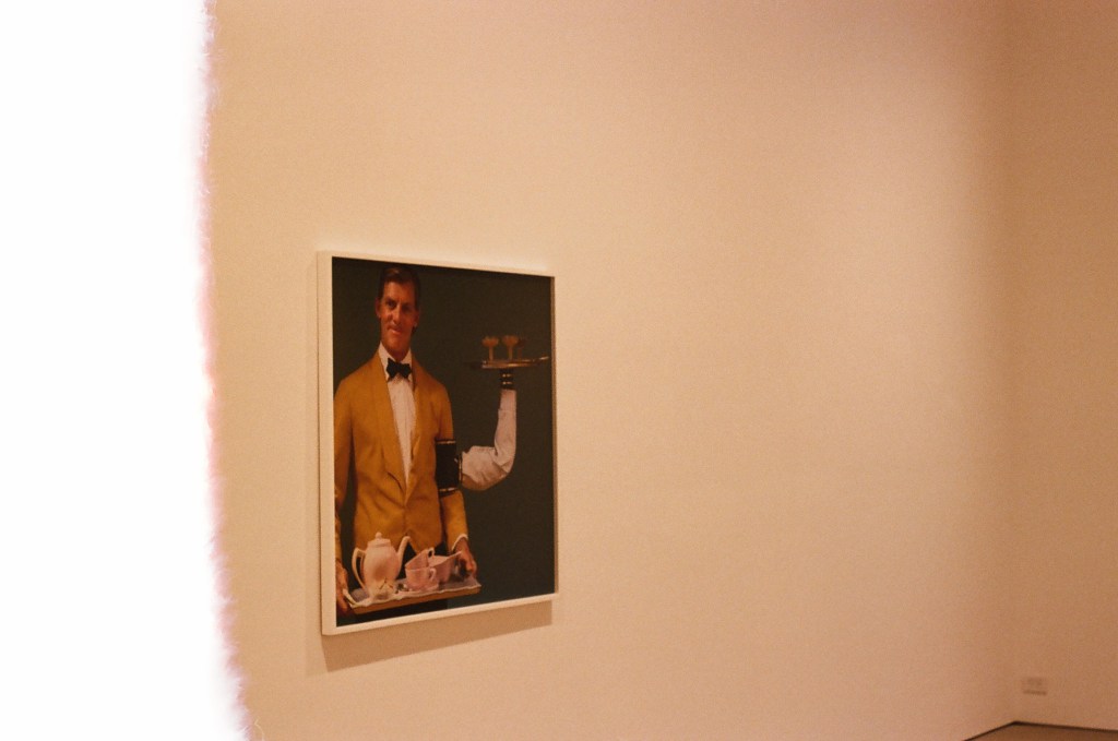 A photo from an exhibition by artist Pippa Garner. The image is of a photograph hanging on a wall, taken at an angle. The photograph is of a white man with a mustache in waitstaff clothing holding a tea set. He has a cuff attached to his arm with another arm holding a drink tray. The image is warm and has a light leak on the left one-quarter.