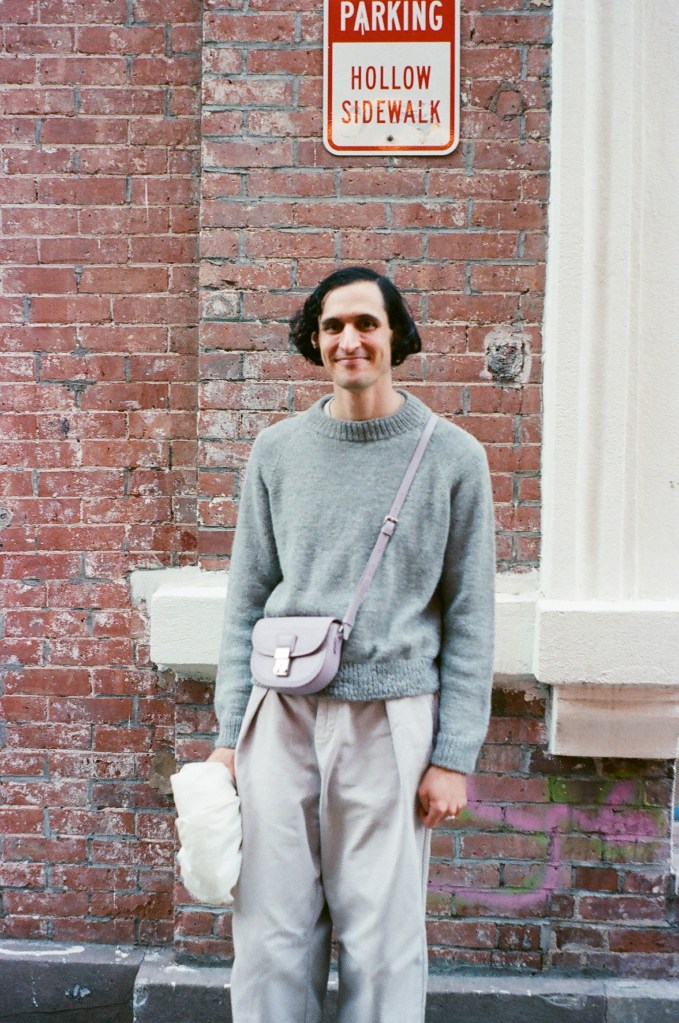 A white man in his early 30s with black hair in a short bob stands in front of a brick wall. He is wearing a gray knit sweater, small lavender cross-body bag, and wide pleated gray pants.