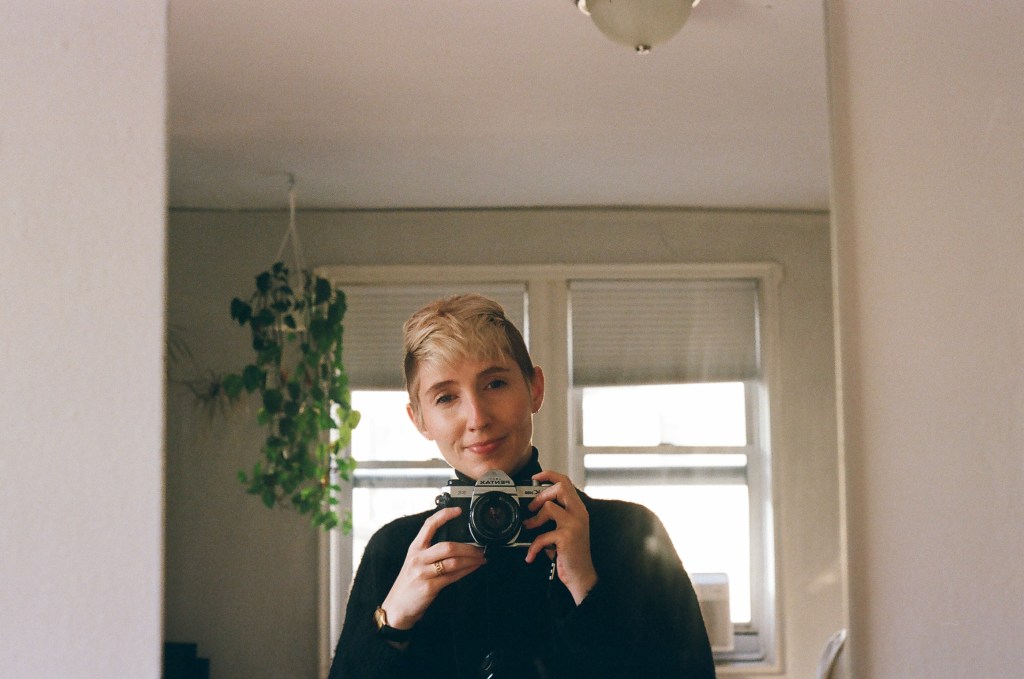 A white woman in her early 30s with short bleached hair taking a selfie in a mirror with a film camera. She is wearing all black and smiling slightly. Behind her, a viney pothos plant hangs from the ceiling next to two windows.