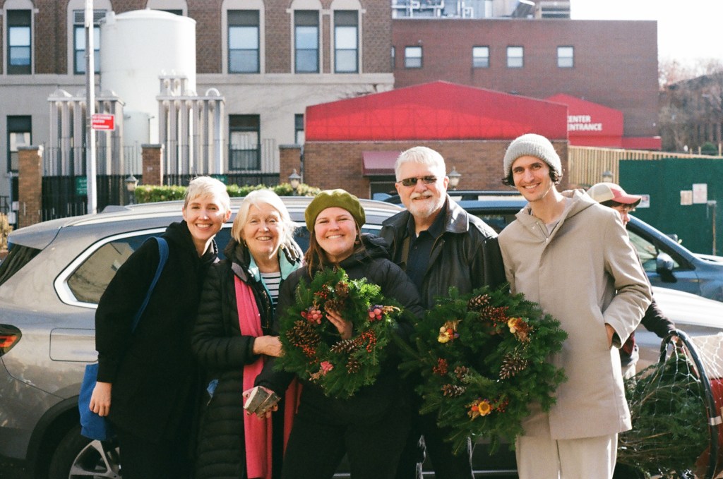Five people stand in front of a car. Two are holding Christmas wreaths. They are dressed in winter clothes but it is also sunny.