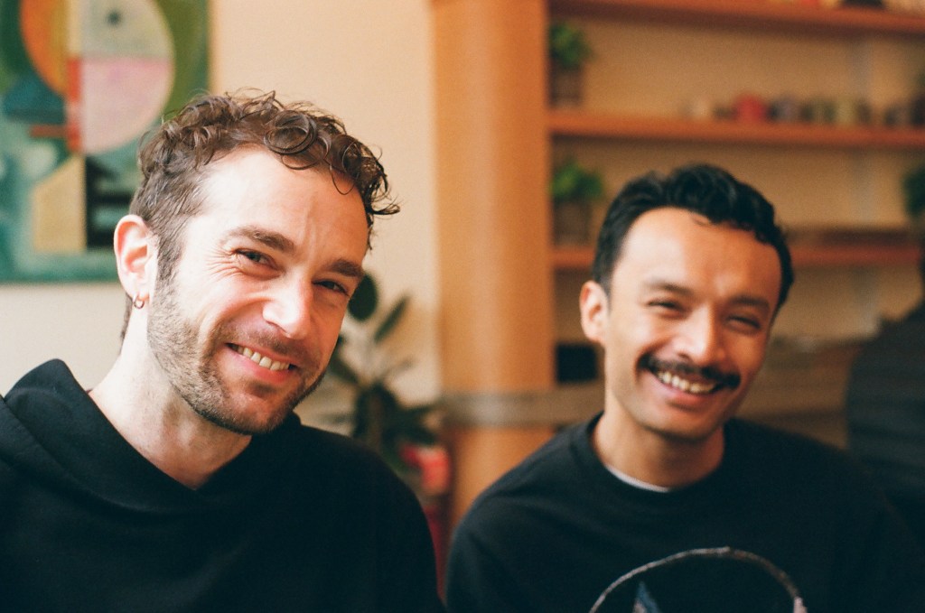 Two men in their late 30s smile at the camera. They are seated. One man is white with short, curly light brown hair, a short beard, and an earring visible to the camera. He is wearing a black hoodie and the light rakes across his face. The second man is seated to his left (right side of image). He is Cambodian, with short wavy black hair and a mustache. He is wearing a black t-shirt. He is slightly out of focus.
