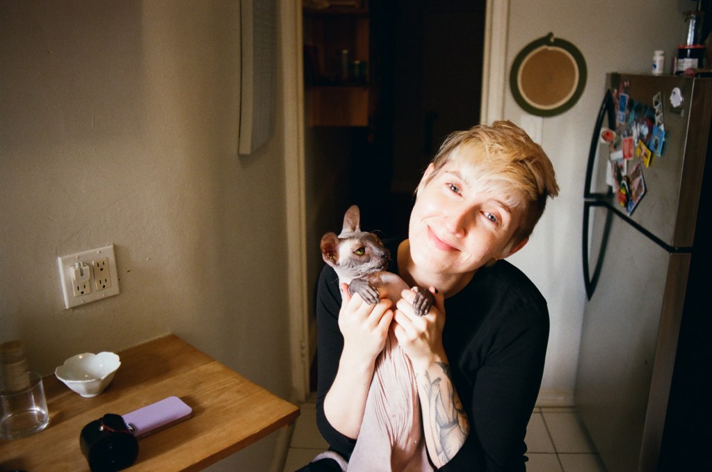 A white woman in her early 30s looks into the camera and smiles in a kitchen. She has short bleached hair. She is holding up a hairless cat on her lap. The cat is light gray with a pink belly. The cat it looking at the woman skeptically.