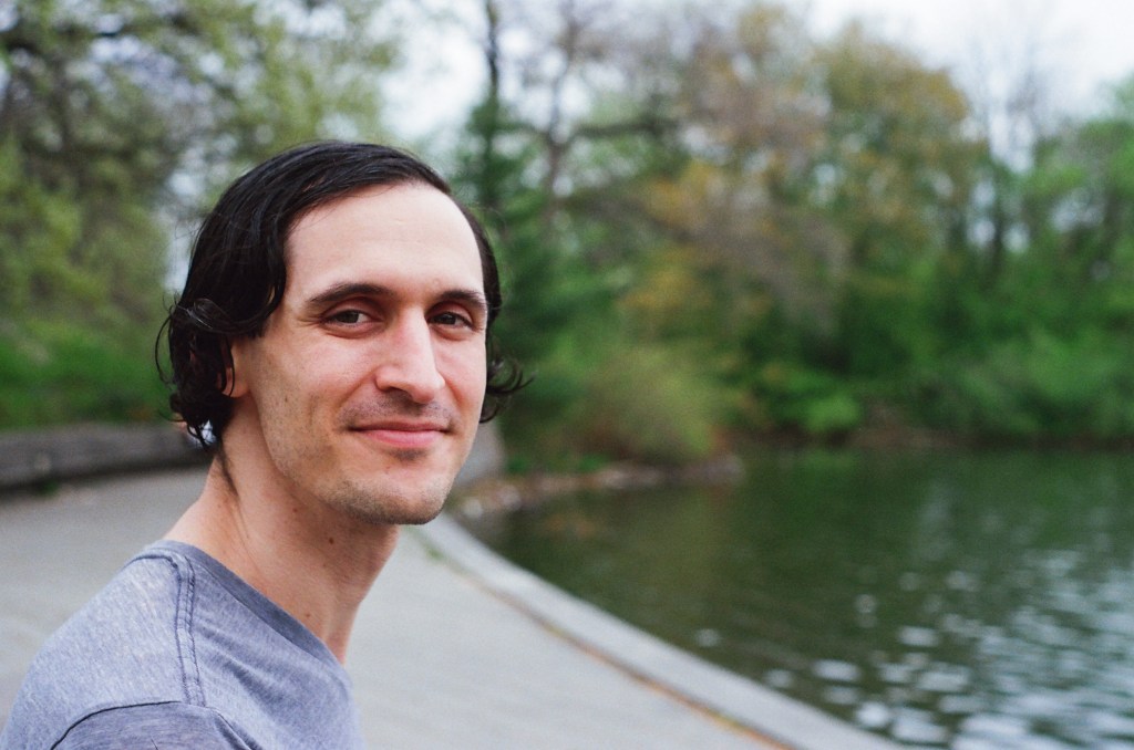 early 30s white man with a wavy black bob is smiling slightly at the camera, with a pond and trees in the background
