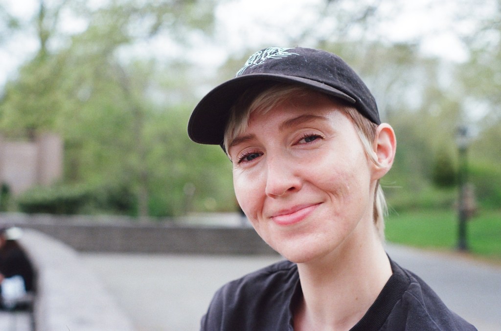 early 30s white woman with short bleached hair, wearing a black baseball hat, smiles at the camera, with blurry trees in the background