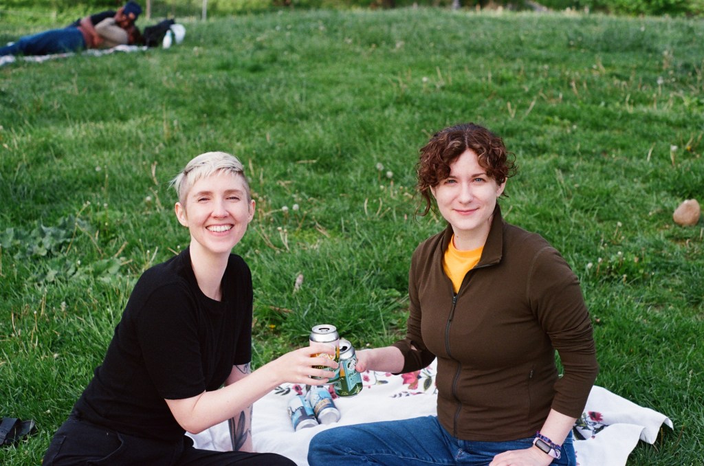 two white women in their early thirties sit on a blanket on very green grass, holding beer cans and smiling at the camera. in the background and out of focus, two people lay on the grass kissing