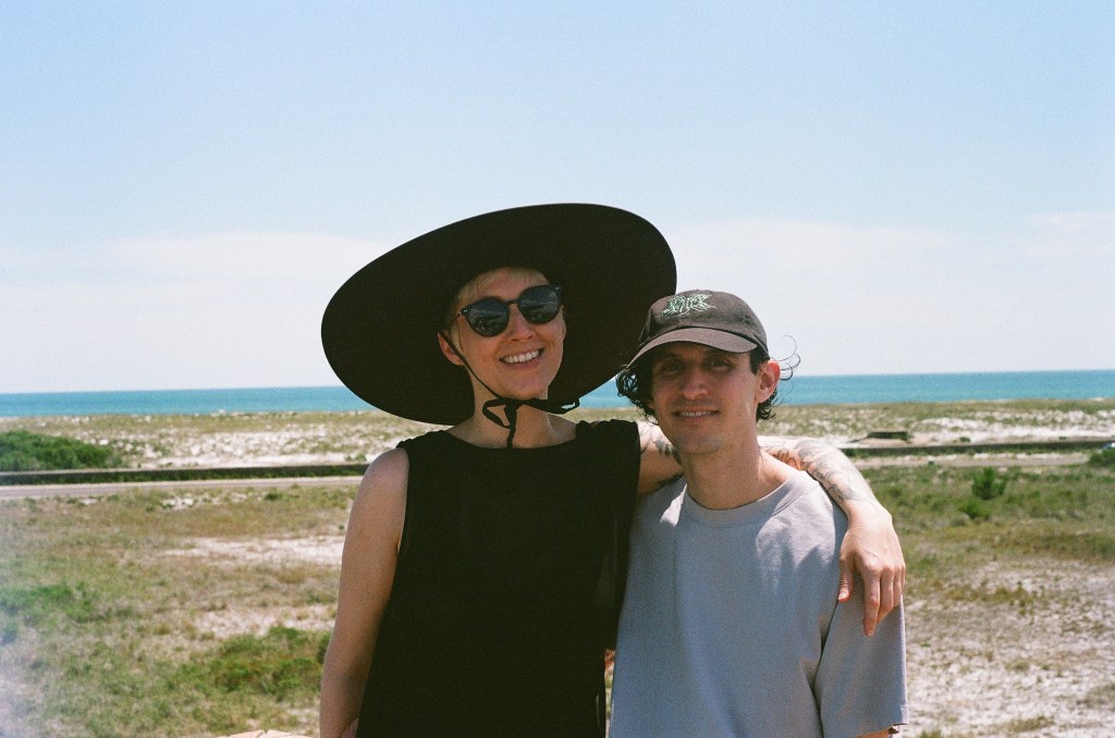Two white people in their early 30s, a woman and a man, smile at the camera. She is on the left, wearing a sleeveless black top, sunglasses, and a wide-brimmed black hat that ties under her chin. He is wearing a short-sleeved light gray shirt and a black baseball hat, under which curly black hair is visible. They are standing in front of a landscape of scrubby grass and sand, blue water in the background.