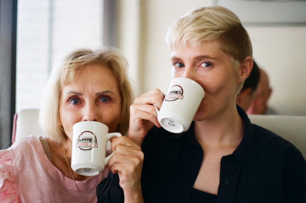Two people pretend to drink from mugs that say "Coffee Cup" with the image of a building printed on them. The person on the left is in her 60s and shorter, with medium length blonde hair and a pink shirt on. The other is taller, with very short blonde and brown hair cut into a soft mohawk-like cut. She is wearing a black button-up shirt with another shirt underneath.