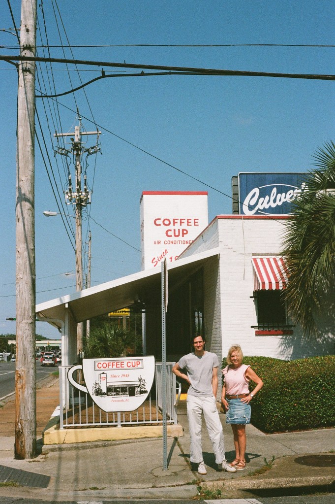 Two people stand next to a stop sign in front of an older white brick building with red accents. The building has a chimney on which is written "Coffee Cup air conditioned." The word "since" is also visible, but not the date. The two people are both white and smile at the camera. One is a tall man in his early thirties wearing light-colored pants and shirt with medium length black hair. The other is a shorter woman in her mid-60s with medium length blonde hair, a denim skirt, and a short-sleeved pink shirt.