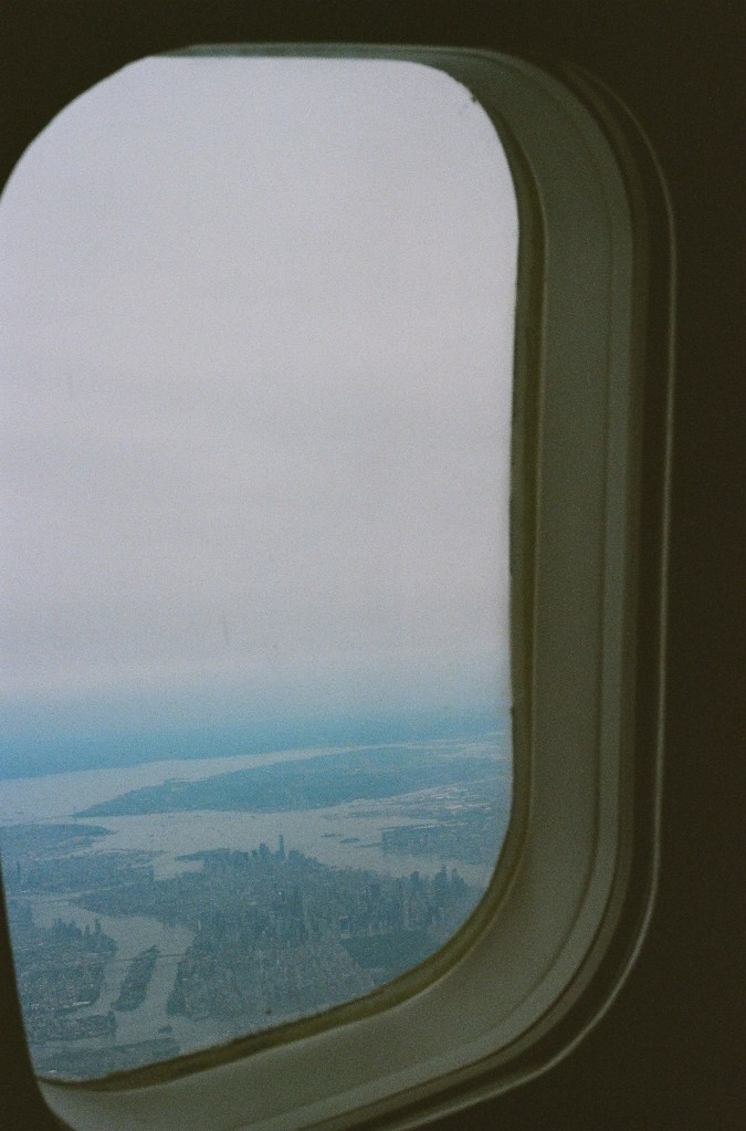 A photo out an airplane window, shortly after take-off from LaGuardia Airport in New York City. Manhattan is seen outside the window, with Roosevelt Island and some of Queens visible. Staten Island is visible in the background without much detail.