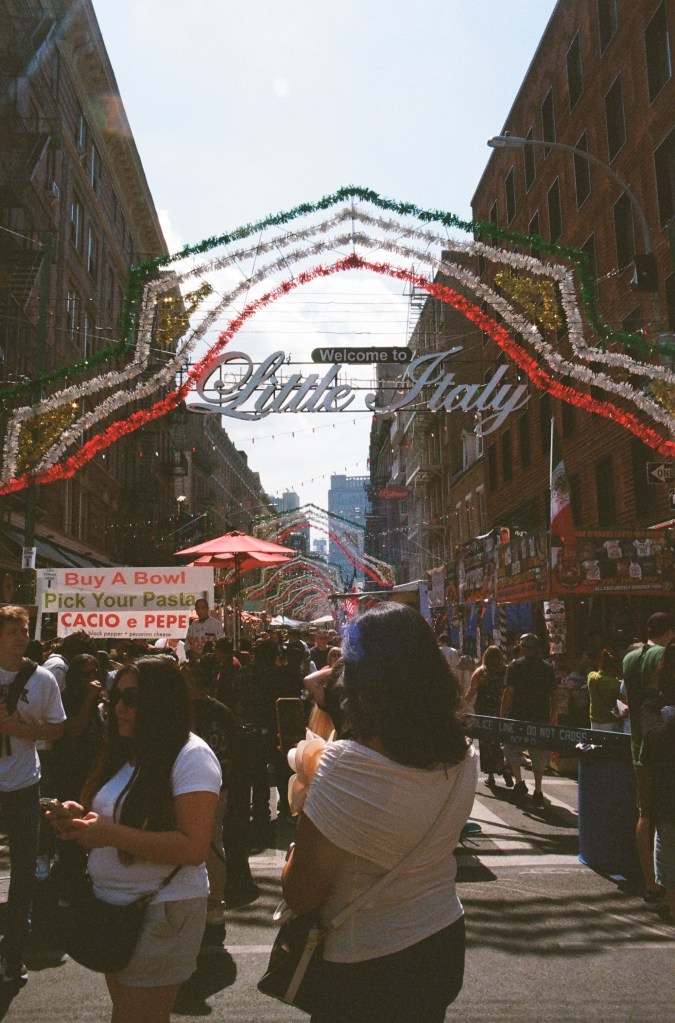 Feast of San Gennaro in Little Italy, with green, white, and red banners across the street and many people in the street. A sign reads "Welcome to Little Italy."
