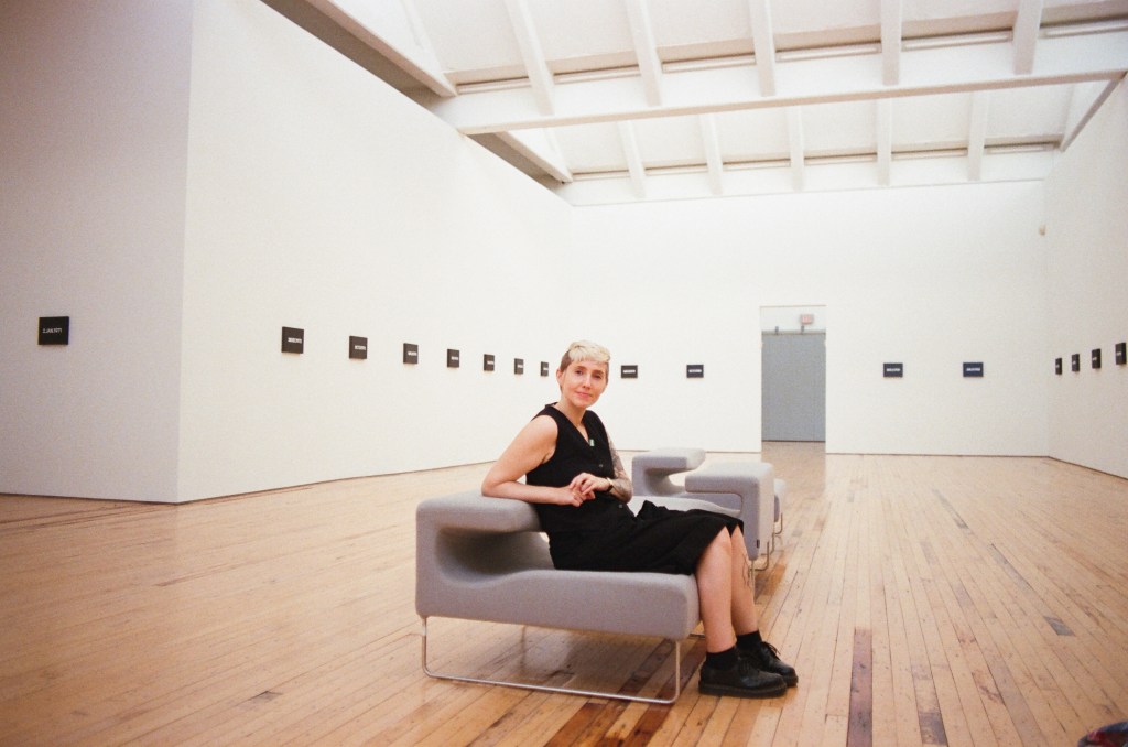 A mid-30s white woman sits back in a gray chair, looking at the camera. She is wearing all black and has short bleach-blonde hair. The room contains  many artworks by On Kawara.