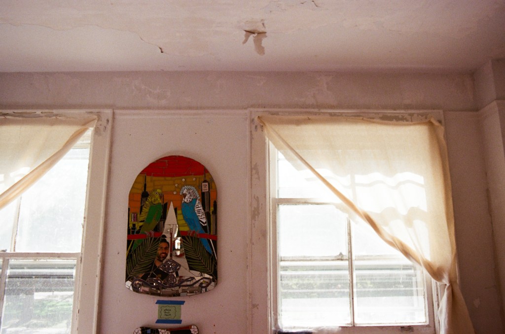 A somewhat dilapidated room with a mosaic by Marco DaSilva hung between two windows. The mosaic has two parakeets and has a reflective surface in the middle, where a man with a beard is reflected (the artist).