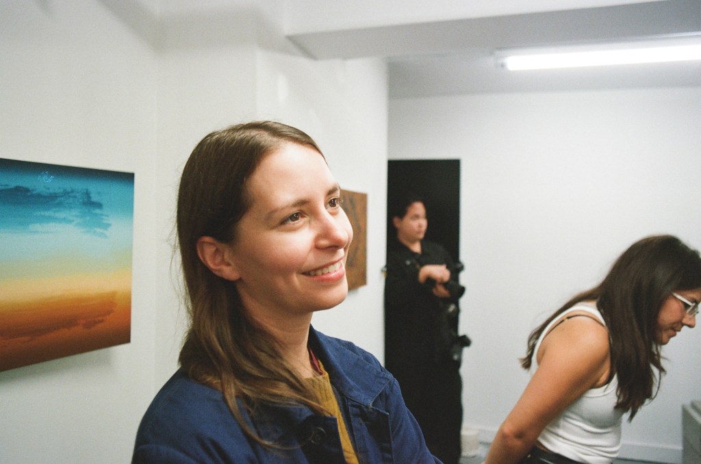 A white woman in her mid-30s with shoulder-length brown hair, a blue jacket, and a golden sweater, smiles at a person not in the frame. She is in a gallery, and part of a painting is seen behind her and to the left. A photographer and another visitor are somewhat blurry in the background.