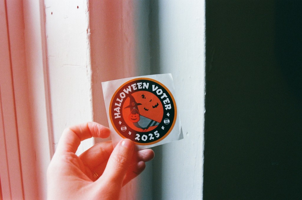 A white person holds a 'Halloween Voter 2025" sticker near the window. There's a light leak across half the photo, making it orange. The voter sticker is a pigeon in a witches hat with a trick-or-treat plastic pumpkin in its beak, with bats in the background.