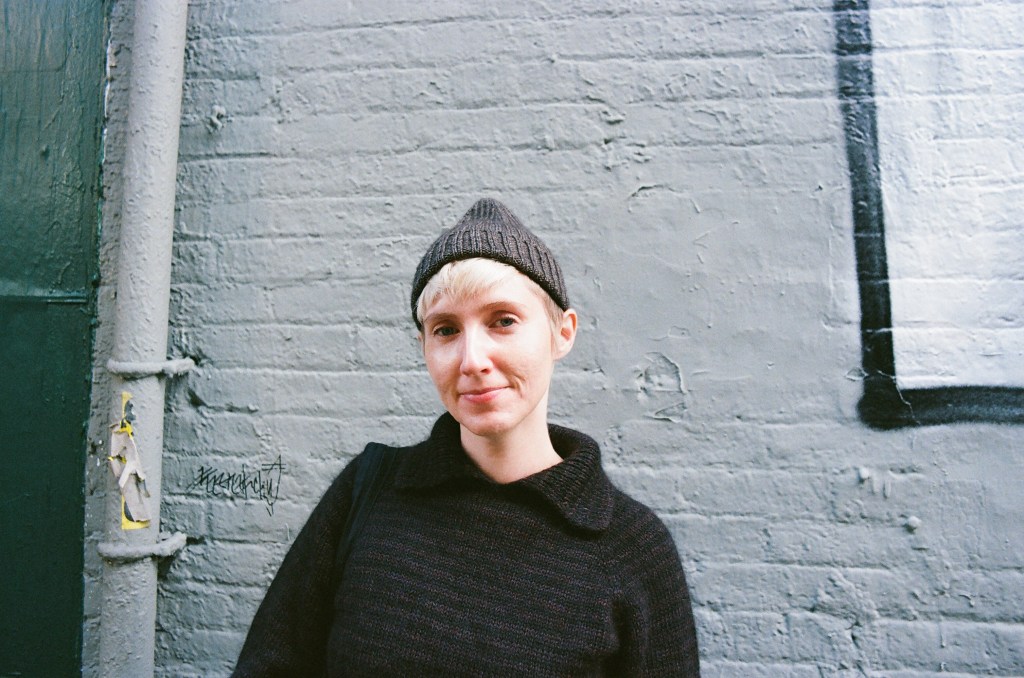 A white woman in her early 30s stands in front of a somewhat distressed white brick wall. She is smiling slightly at the camera, wearing a brown pointy beanie above her ears and a striped collared sweater.