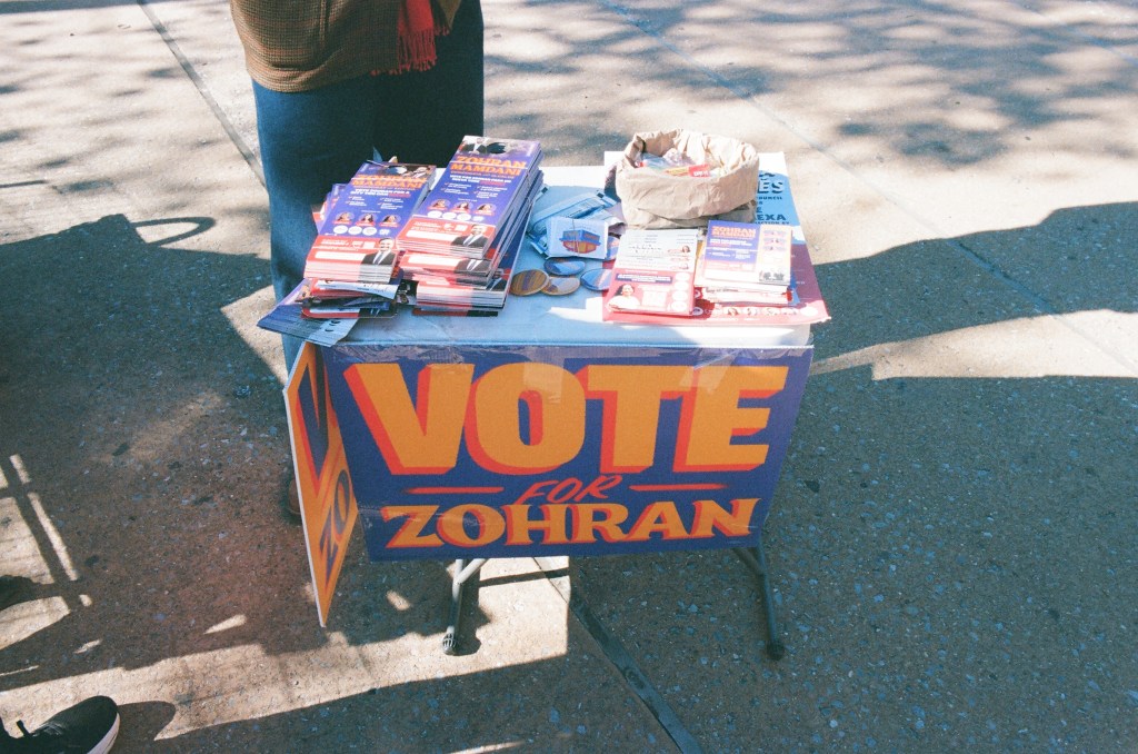 A canvassing table for Zohran Mamdani, with a "Vote for Zohran" sign hanging from its front. Its top is covered in flyers, stickers, and buttons. 