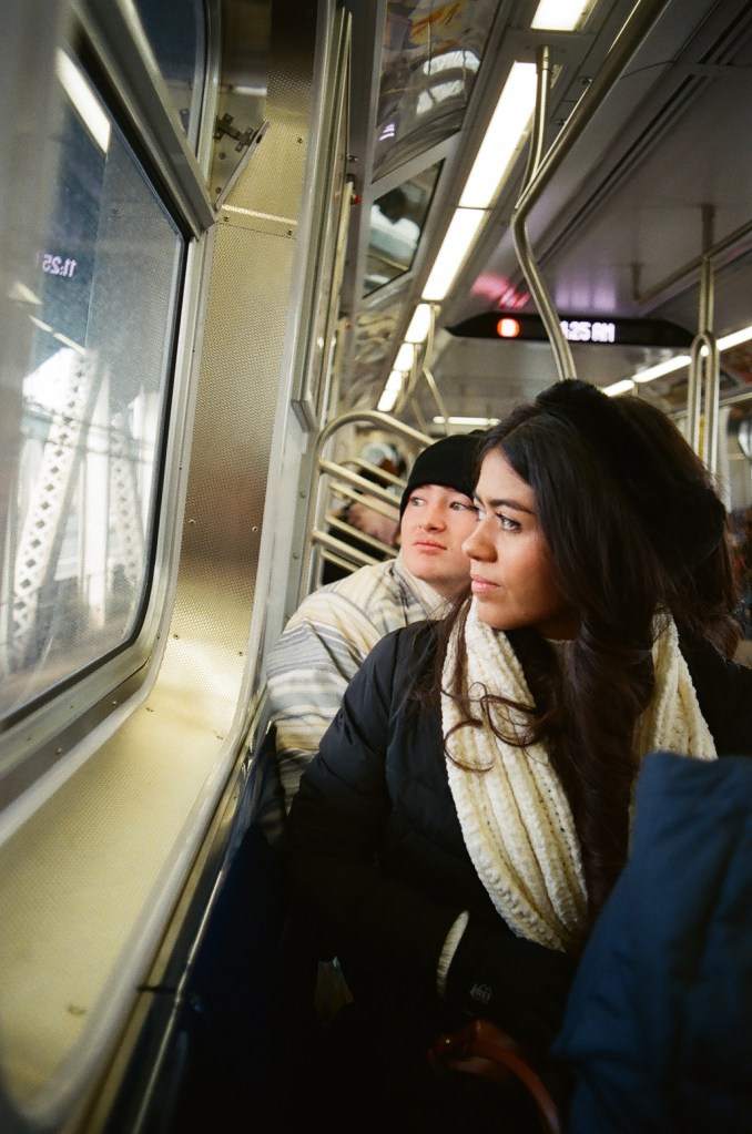 Two people in their early 20s, one a Latina woman and one a white man, look out the window of a subway car.