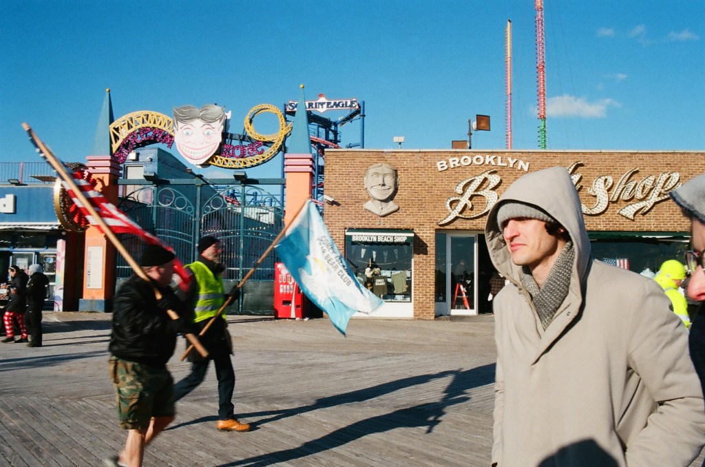 Coney Island boardwalk on New Year's Day. Two white men carry flags from left to right. One flag is for the Coney Island Polar Bear Club. Another white man bundled against the cold in a gray coat, gray scarf, and hat, crosses right to left.