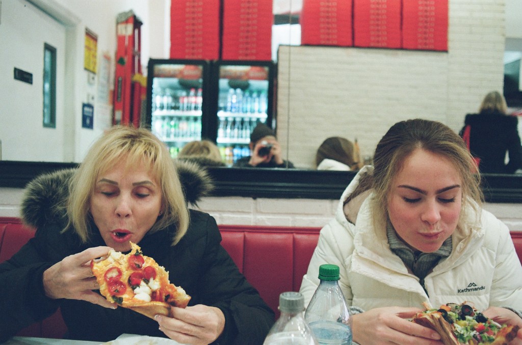 Two white women in winter coats, one in her 60s and the other in her 20s, focus on eating pizza. A reflective mirror behind them shows the photographer in a winter hat.