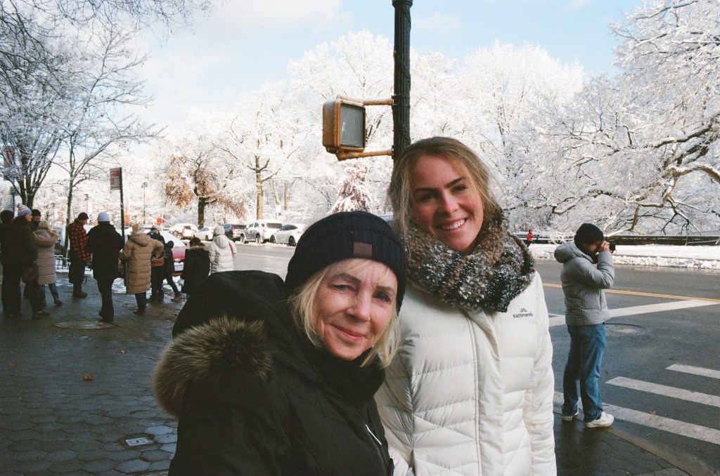Two white women in winter coats, one in her 60s and the other in her 20s, smile at the camera on the corner across from Central Park, covered in fresh snow.