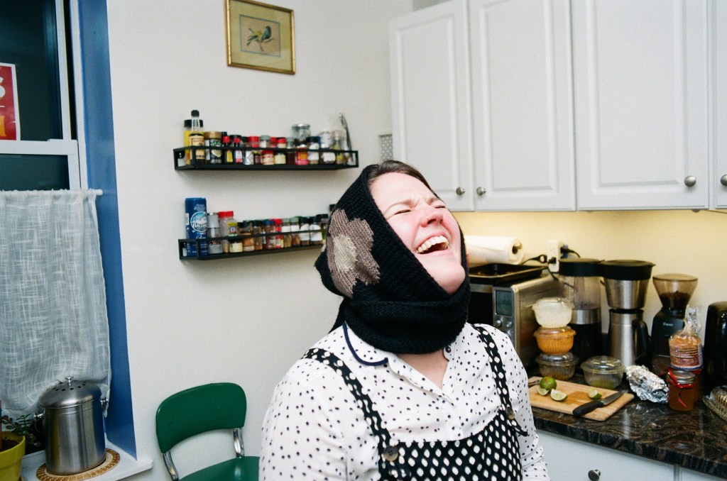 A white woman in her late 30s wearing a knit headwrap with a flower on it and polka-dotted clothes laughs in a kitchen with her head thrown back.