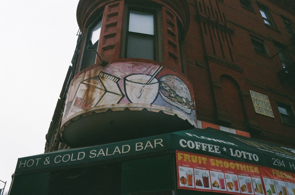 An old handpainted sign above a bodega at the bottom of an older brick building in Park Slope, Brooklyn. The visible portion of the sign has milk, a burger, and a beverage with a straw.
