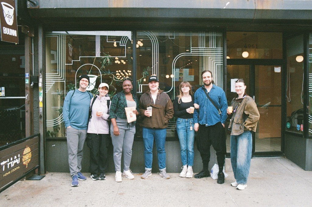 A group of seven people, four women and three men, stand in front of a coffee shop and smile at the camera.