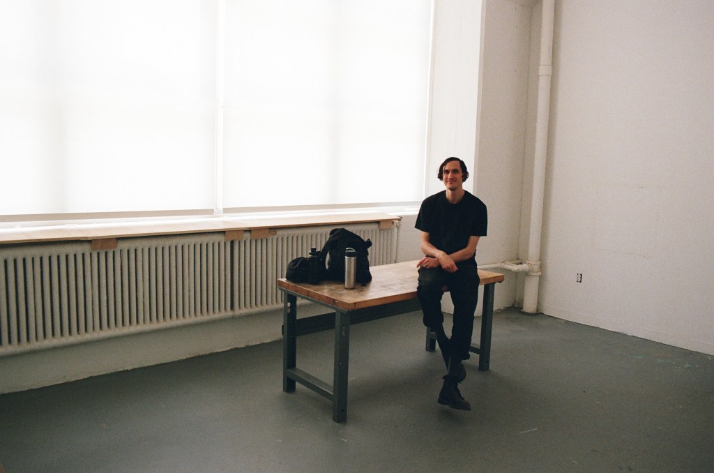 A white man wearing all black sits on a table in an empty art studio, with filtered light coming through windows behind him.