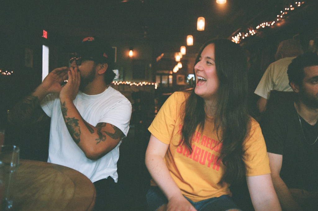 Two people, a man and a woman, look to the left with big smiles, as if yelling. They are at a bar that has hung fairy lights around.